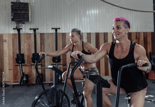 Two muscular athletic women exercising on air bikes at the gym. Functional training cross class working out