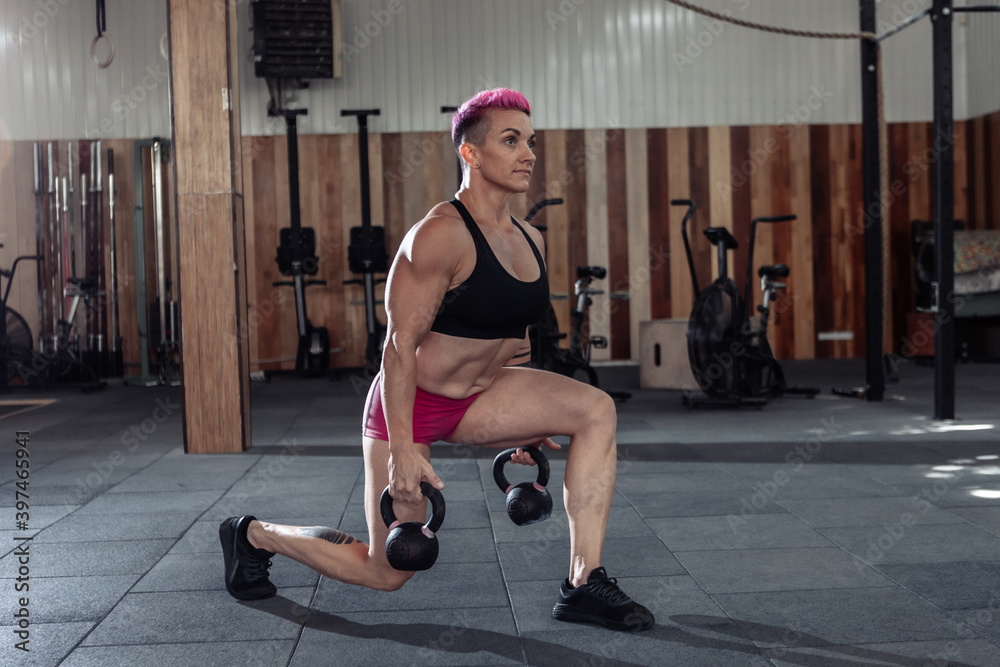 Fototapeta premium Muscular extraordinary female athlete with short pink hair trains lunges with heavy kettlebells. Functional, cross training in modern gym.