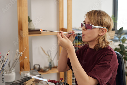 Young skilled female lampworking master blowing into long tube by workplace photo