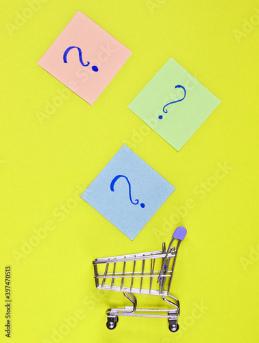 Shopping cart with question marks on memo pieces of paper against green background. Minimalistic shopping concept. Top view