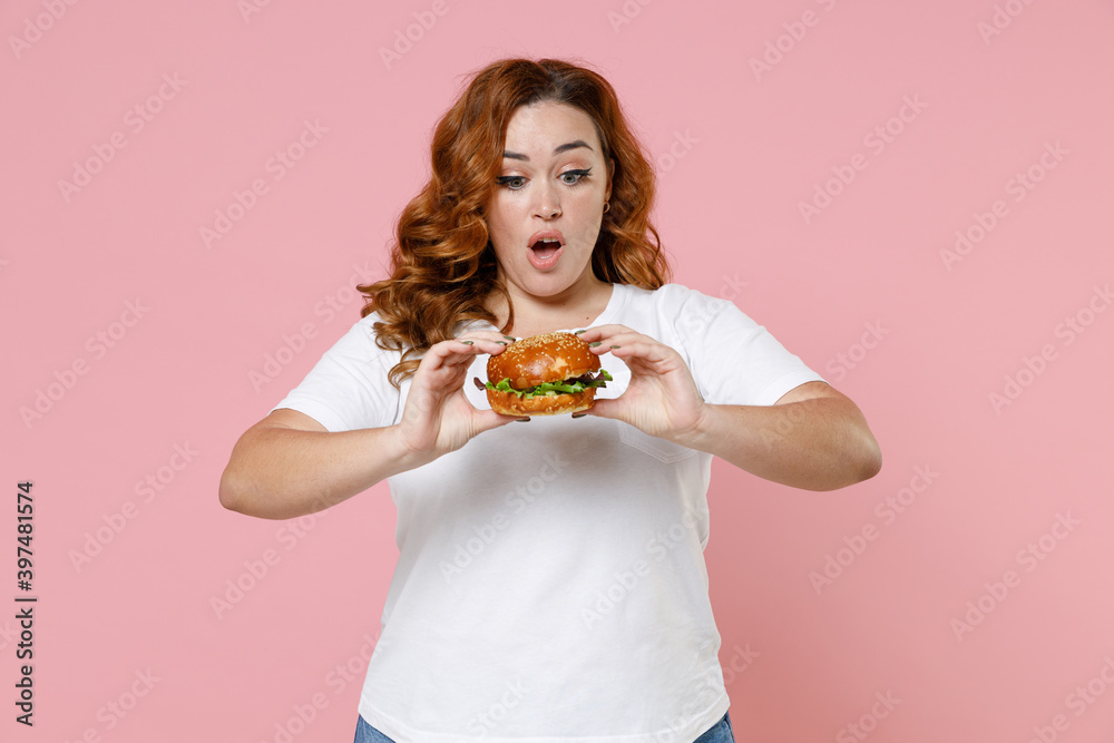 Perplexed young redhead plus size body positive woman 20s in white casual t-shirt hold eating american classic fast food burger keeping mouth open isolated on pastel pink background studio portrait. Perplexed young redhead plus size body positive woman 20s in white casual t-shirt hold eating american classic fast food burger keeping mouth open isolated on pastel pink background studio portrait.