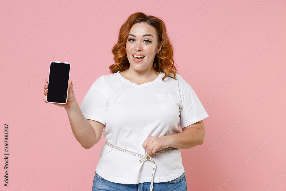 Funny young redhead plus size body positive woman 20s in white t-shirt hold mobile cell phone with blank empty screen measuring waist with measure tape isolated on pink background studio portrait. Funny young redhead plus size body positive woman 20s in white t-shirt hold mobile cell phone with blank empty screen measuring waist with measure tape isolated on pink background studio portrait.
