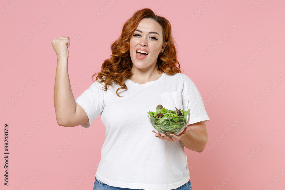 Joyful young redhead plus size body positive woman 20s wearing white casual t-shirt hold vegetable salad in glass bowl doing winner gesture isolated on pastel pink color background studio portrait. Joyful young redhead plus size body positive woman 20s wearing white casual t-shirt hold vegetable salad in glass bowl doing winner gesture isolated on pastel pink color background studio portrait.