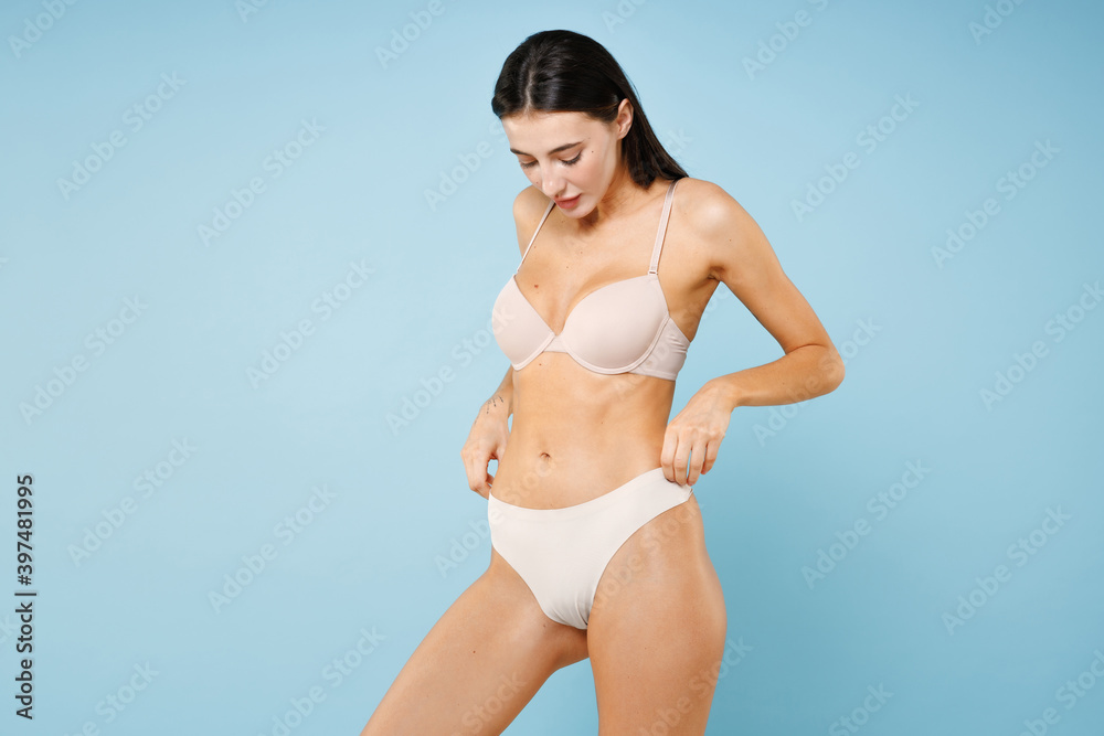 Beautiful attractive sexy seductive gorgeous young brunette woman 20s in beige underwear showing fit body standing posing looking down isolated on pastel blue colour wall background, studio portrait. Beautiful attractive sexy seductive gorgeous young brunette woman 20s in beige underwear showing fit body standing posing looking down isolated on pastel blue colour wall background, studio portrait.