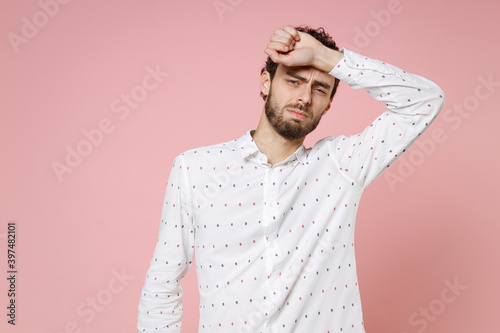 Dissatisfied exhausted tired young bearded man 20s wearing basic casual white shirt standing put hand on head looking camera isolated on pastel pink color wall background studio portrait. photo