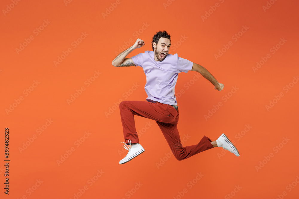Full length side view of screaming young bearded man 20s in casual violet t-shirt jumping like running keeping mouth open looking camera isolated on bright orange color background studio portrait.
