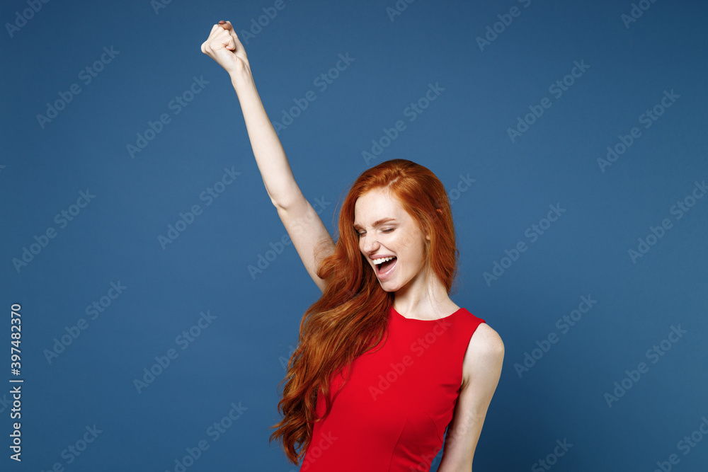 Joyful happy young redhead woman 20s wearing bright red elegant evening dress standing doing winner gesture clenching fists keeping eyes closed isolated on blue color wall background studio portrait.