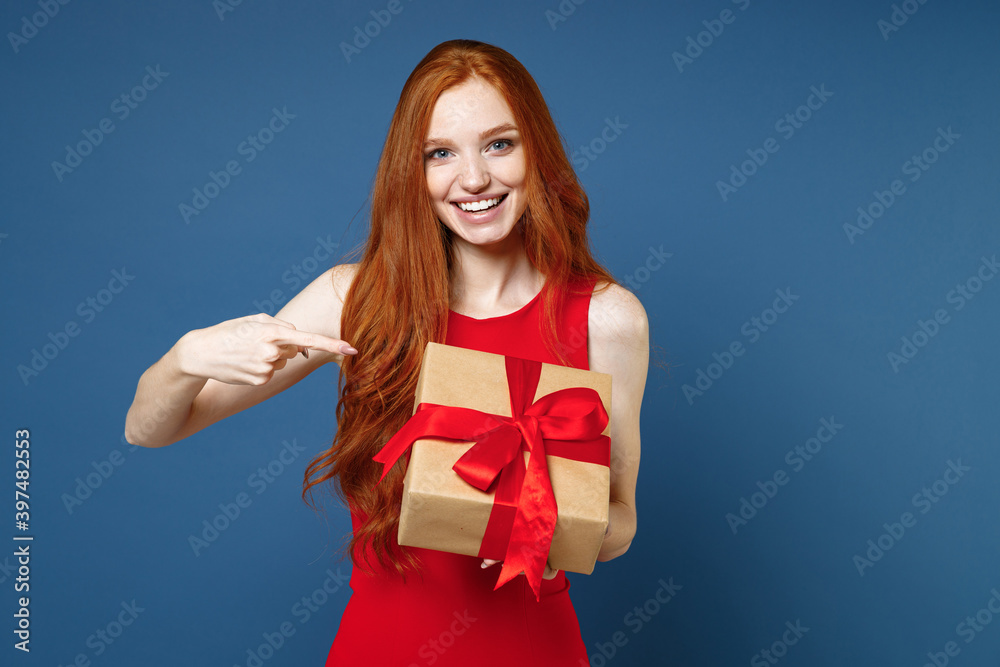 Funny young redhead woman in red elegant evening dress pointing index finger on present box with gift ribbon bow isolated on blue background. St. Valentine's Day Women's Day birthday, holiday concept. Funny young redhead woman in red elegant evening dress pointing index finger on present box with gift ribbon bow isolated on blue background. St. Valentine's Day Women's Day birthday, holiday concept.