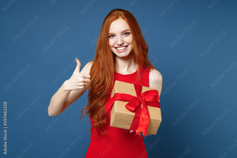Smiling young redhead woman in red elegant evening dress hold present box with gift ribbon bow showing thumb up isolated on blue background. St. Valentine's Day Women's Day birthday holiday concept. Smiling young redhead woman in red elegant evening dress hold present box with gift ribbon bow showing thumb up isolated on blue background. St. Valentine's Day Women's Day birthday holiday concept.