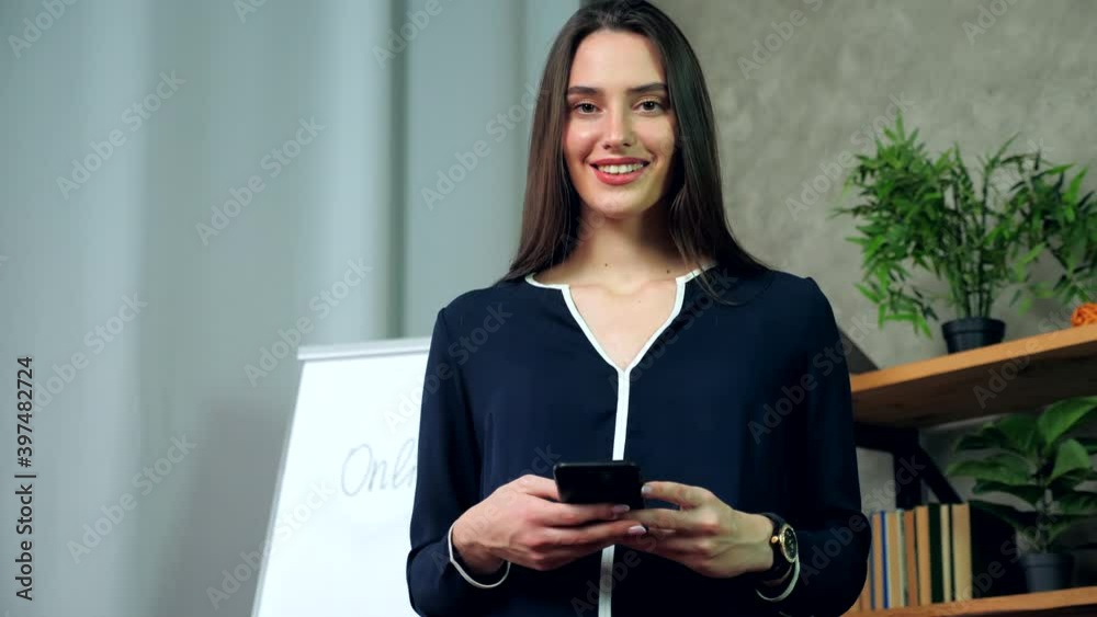 Beautiful smiling teacher coach holding phone in hands looking camera in office. Positive business woman trainer tutor uses smartphone Beautiful smiling teacher coach holding phone in hands looking camera in office. Positive business woman trainer tutor uses smartphone