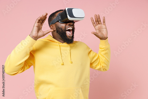 Excited young african american man 20s in casual yellow streetwear hoodie watching in vr headset gadget rising spreading hands looking aside isolated on pastel pink color background studio portrait.