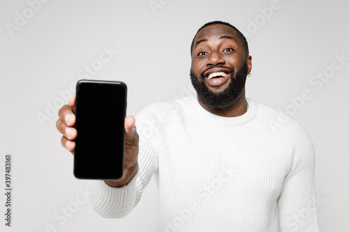 Cheerful funny young african american man 20s wearing casual basic sweater hold mobile cell phone with blank empty screen mock up copy space isolated on white color wall background studio portrait.