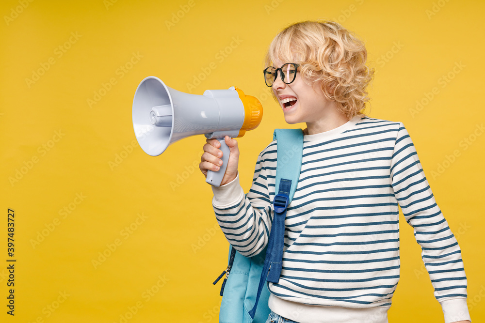 Irritated male kid teen boy 10s years old in striped sweatshirt eyeglasses backpack screaming in megaphone looking aside isolated on yellow color background, child studio portrait. Education concept. Irritated male kid teen boy 10s years old in striped sweatshirt eyeglasses backpack screaming in megaphone looking aside isolated on yellow color background, child studio portrait. Education concept.