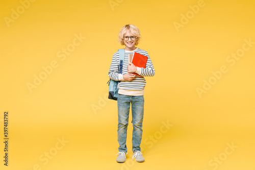 Full length of funny little male kid teen boy 10s years old in striped sweatshirt eyeglasses backpack holding school books isolated on yellow color background child studio portrait. Education concept.