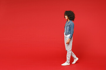 Full length side view of smiling little african american kid girl 12-13 years old in casual denim jacket looking aside isolated on red background children studio portrait. Childhood lifestyle concept.
