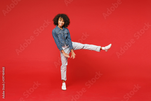 Full length of funny little african american kid girl 12-13 years old in casual denim jacket dancing rising leg isolated on bright red background children studio portrait. Childhood lifestyle concept.