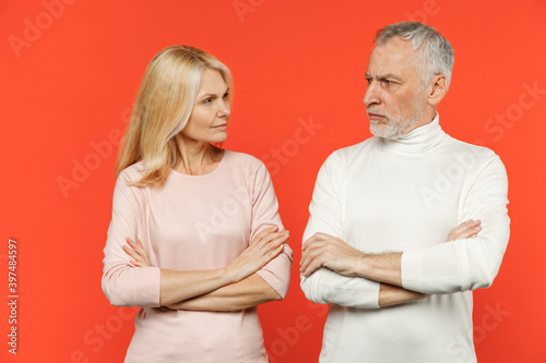 Displeased couple two friends elderly gray-haired man blonde woman in white pink casual clothes standing holding hands crossed looking at each other isolated on orange wall background studio portrait.