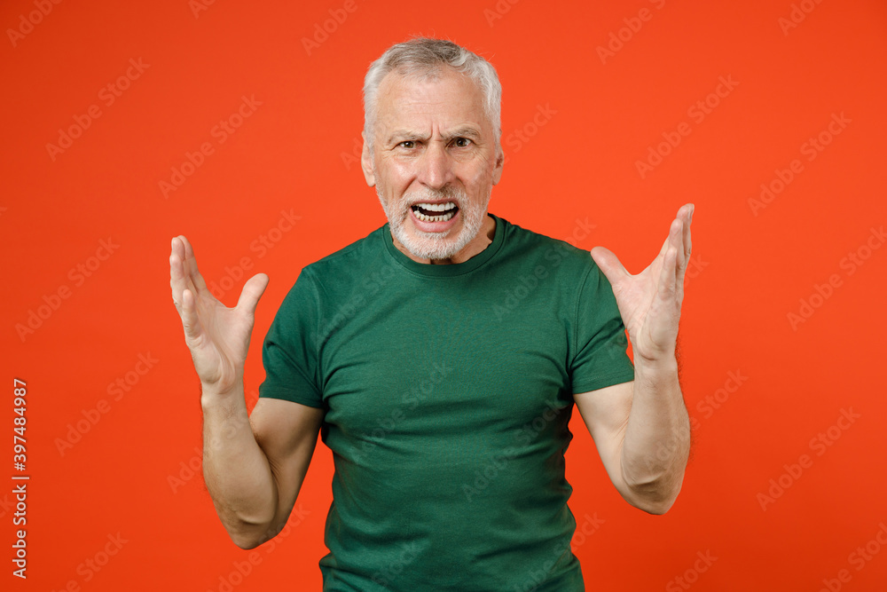 Angry irritated elderly gray-haired mustache bearded man in basic green t-shirt standing spreading hands screaming swearing looking camera isolated on bright orange color background studio portrait. Angry irritated elderly gray-haired mustache bearded man in basic green t-shirt standing spreading hands screaming swearing looking camera isolated on bright orange color background studio portrait.