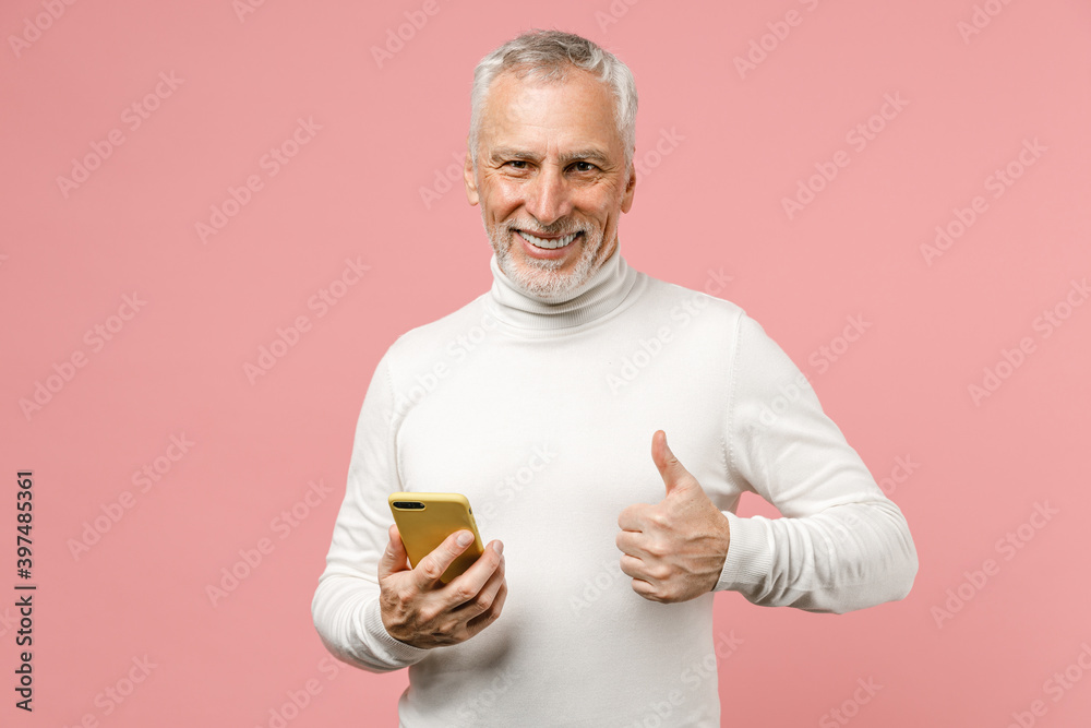 Smiling elderly gray-haired mustache bearded man in casual white turtleneck standing showing thumb up using mobile cell phone typing sms message isolated on pastel pink background studio portrait. Smiling elderly gray-haired mustache bearded man in casual white turtleneck standing showing thumb up using mobile cell phone typing sms message isolated on pastel pink background studio portrait.