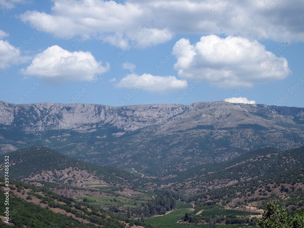 View of a valley of mountains and hills covered with a forest of low trees. Lush clouds float across the blue sky.