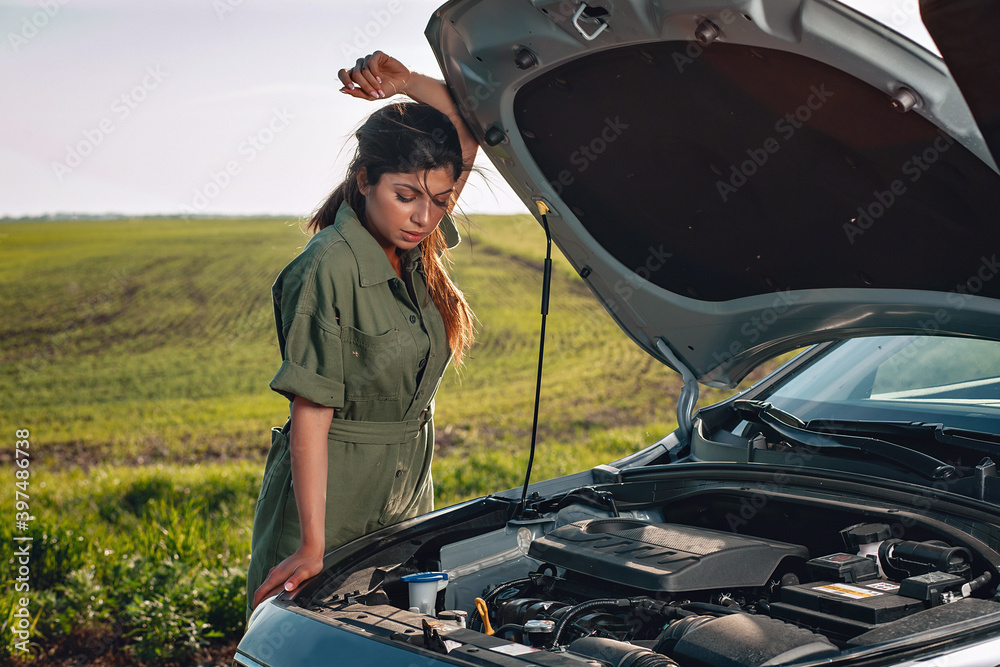 brunette slim girl opened the hood of her car, leaning on the door and looking down in the middle of the field