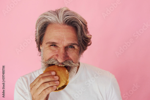 Eats delicious burger. Stylish modern senior man with gray hair and beard is indoors