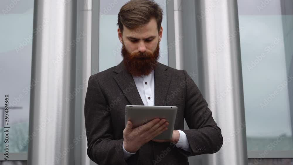 Modern Young bearded Business man working with a digital tablet. Young hipster businessman holding tablet in hands outdoor. Working online with a tablet while standing outside on an office building. Modern Young bearded Business man working with a digital tablet. Young hipster businessman holding tablet in hands outdoor. Working online with a tablet while standing outside on an office building.