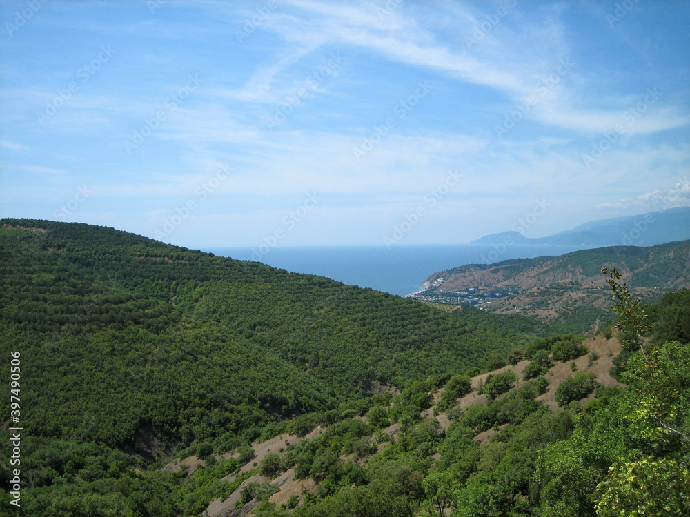 A valley covered with dense low-growing forest and shrubs descends to the blue sea coast. Clouds float in the high sky.
