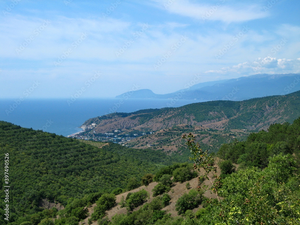 A valley covered with dense low-growing forest and shrubs descends to the blue sea coast. Clouds float in the high sky.