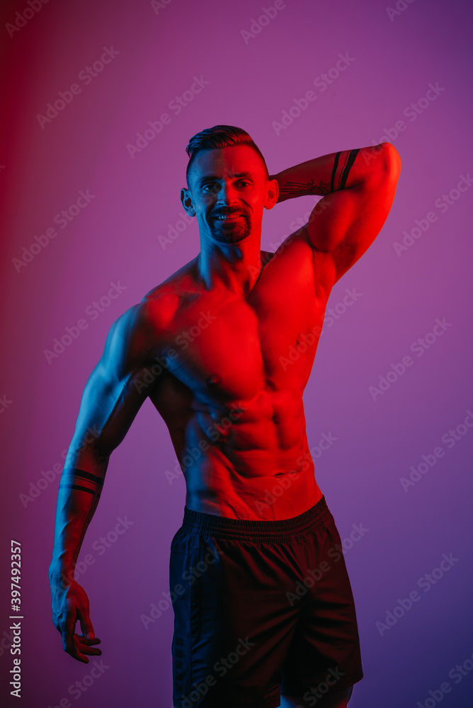 A muscular man with a beard is posing with an arm behind his head under the blue and red light. An athletic guy is demonstrating his sporty torso. An athlete in shorts after a workout. A muscular man with a beard is posing with an arm behind his head under the blue and red light. An athletic guy is demonstrating his sporty torso. An athlete in shorts after a workout.