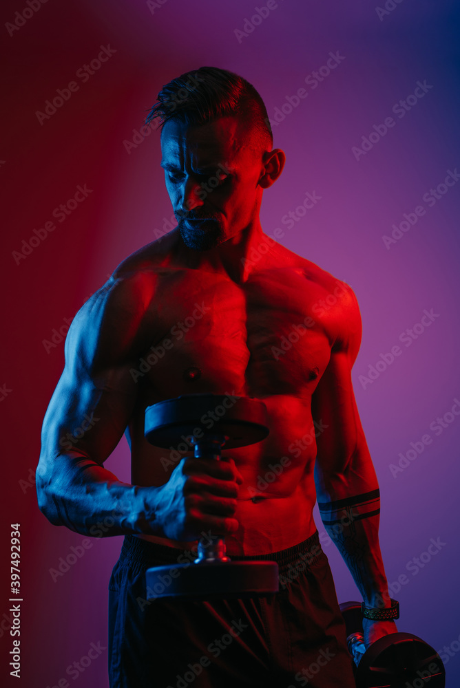 A muscular man with a beard who is doing bicep hammer curls with dumbbells under blue and red lights. An athletic guy is demonstrating his sporty physique. An athlete is training his arms hard.