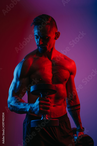Obraz na płótnie A muscular man with a beard who is doing bicep hammer curls with dumbbells under blue and red lights. An athletic guy is demonstrating his sporty physique. An athlete is training his arms hard. w salonie