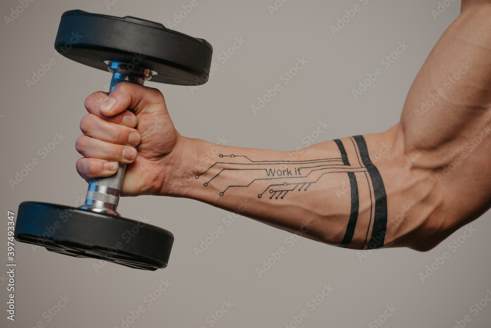 A close photo of a muscular arm which is doing bicep hummer curls with a dumbbell. A forearm of a man with a tattoo: Work it.