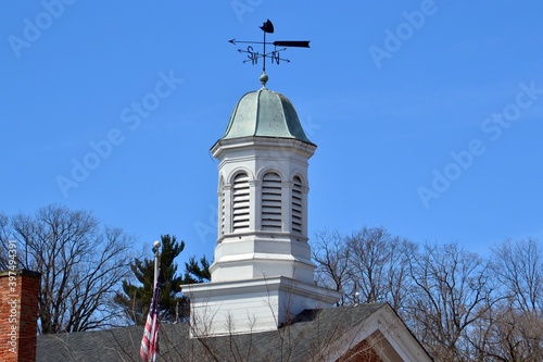 White wooden cupola with a weathervane/wind compass on top sits on a colonial brick roof against a bright blue sky.