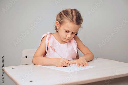 Portrait of a concentrated adorable child in a dress sitting at a white table and writing numbers in a special sheet of paper. Study concept. photo with noise