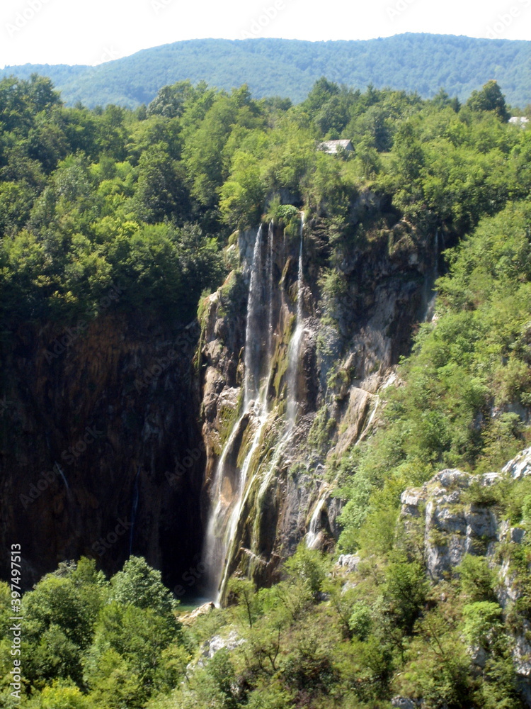 High waterfall in a dense forest. Numerous streams of water fall from the sheer rock face. The waterfall is surrounded by a dense green forest.