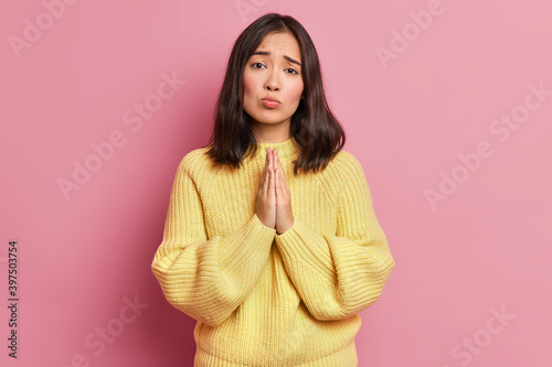 Hopeless brunette woman keeps palms pressed together and looks with imploring expression begs for favor asks to give one more chance wears long sleeved yellow jumper isolated over pink wall. © wayhome.studio 