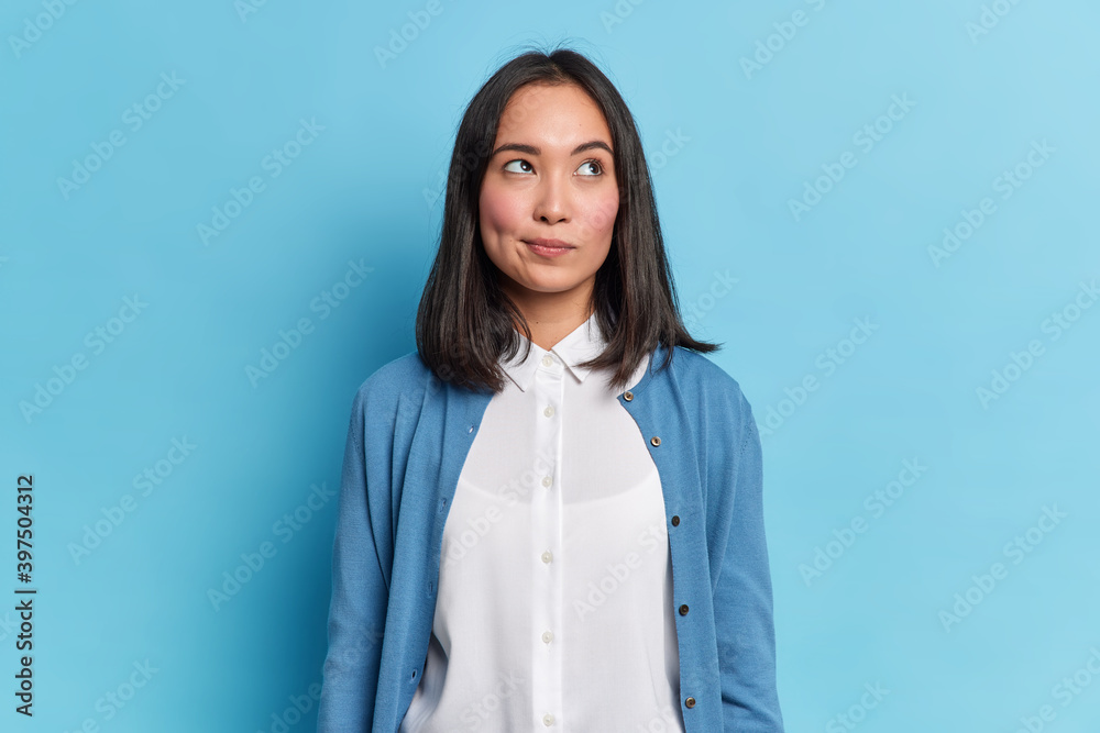 Lovely brunette young woman thinks seriously about something concentrated above has thoughtful expression wears white shirt and long sleeved jumper has rouge cheeks isolated on blue studio wall - obrazy, fototapety, plakaty 