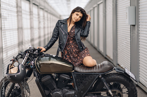Portrait of a sexy adorable pretty woman in a black leather jacket and dress posing next to a black retro motorcycle and looking playfully straight into the camera. Sexuality concept