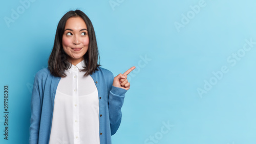 Look at this advert. Pleased brunette young Asian woman points aside shows blank copy space for commercial text promo idea presentation poses against blue background. Attention to nice product photo