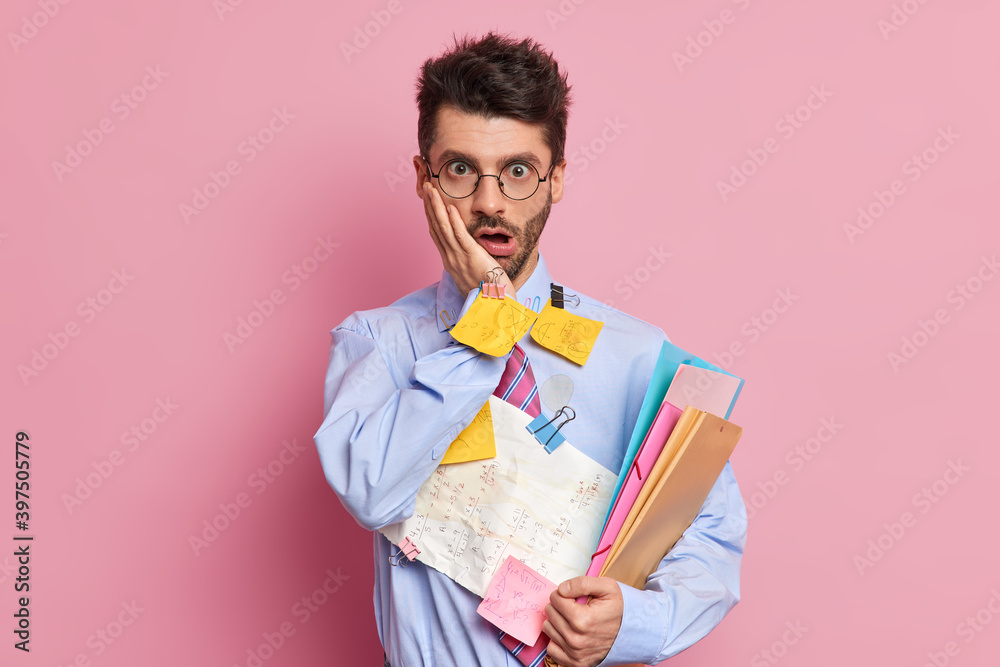 Shocked unshaven man gazes surprisingly keeps hand on cheek holds paper documents with written information data wears formal clothes isolated over pink background has deadline for organizing job Shocked unshaven man gazes surprisingly keeps hand on cheek holds paper documents with written information data wears formal clothes isolated over pink background has deadline for organizing job