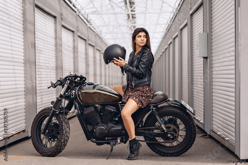 Girl on a motorcycle. Beautiful female driver in a leather jacket and dress sitting on her retro motorcycle and holding a helmet in her hands is getting ready to go. Moto ​​concept