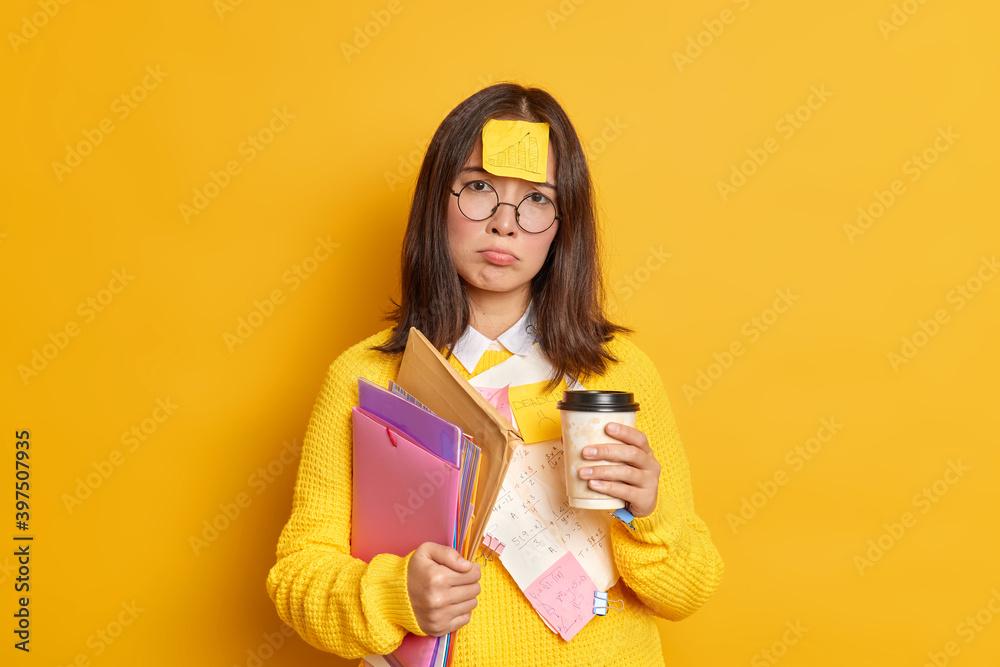 Disappointed tired female graduate prepares for examination session has deadline surrounded by sticky notes and papers holds disposable cup of coffee has unhappy displeased expression stands indoor Disappointed tired female graduate prepares for examination session has deadline surrounded by sticky notes and papers holds disposable cup of coffee has unhappy displeased expression stands indoor