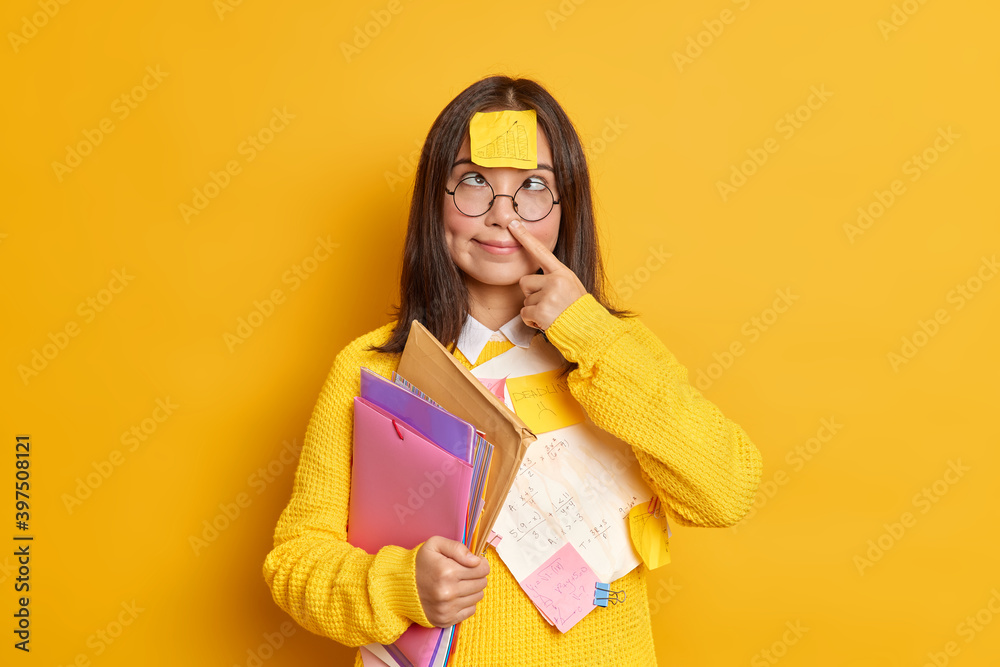 Crazy brunette female student makes fool has funny expression finger stuck in nose wears sticker with graphic on forehead being obssesed with work holds folders prepares for exams isolated on yellow Crazy brunette female student makes fool has funny expression finger stuck in nose wears sticker with graphic on forehead being obssesed with work holds folders prepares for exams isolated on yellow
