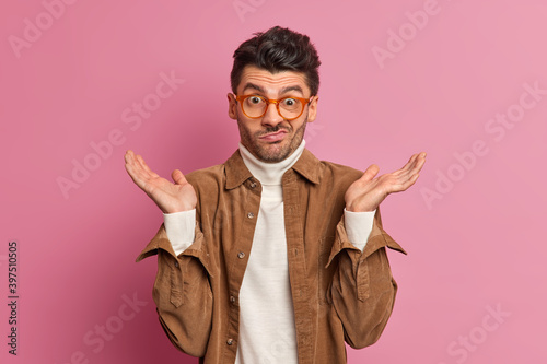 Hesitant clueless unshaven man spreads palms and faces difficult choice looks confused wears transparent glasses and brown shirt poses against rosy background. Uncertain confused European guy photo