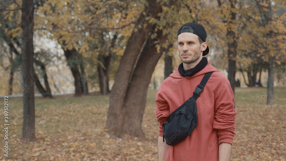 Man runner put on earbuds getting ready for run in a city park. Runner in red hoodie wearing black cap, getting ready for jogging, middle gimbal shot