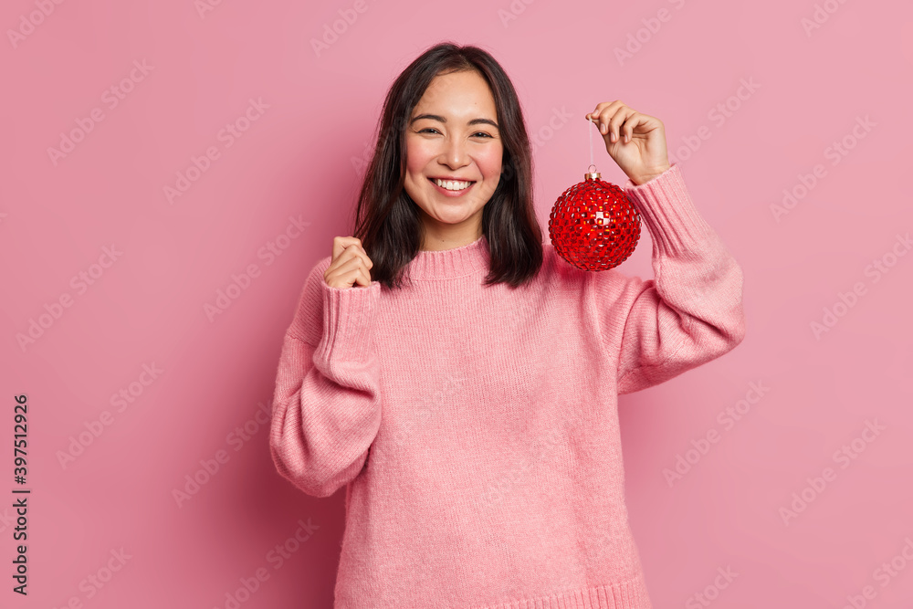 Photo of pretty brunette Asain woman happy to celebrate winter holidays shows best offer for your Christmas tree holds big red ball to decorate prepares for New Year wears casual pink jumper Photo of pretty brunette Asain woman happy to celebrate winter holidays shows best offer for your Christmas tree holds big red ball to decorate prepares for New Year wears casual pink jumper