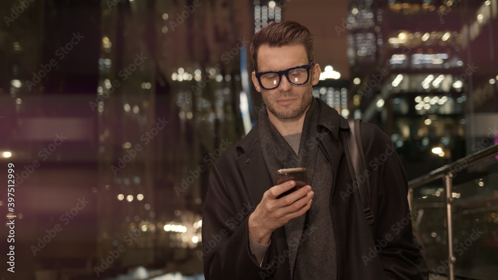 Man in eyeglasses with phone in hand on background of building with lights on. Handheld night shot of caucasian man texting in phone near business building with cars Man in eyeglasses with phone in hand on background of building with lights on. Handheld night shot of caucasian man texting in phone near business building with cars