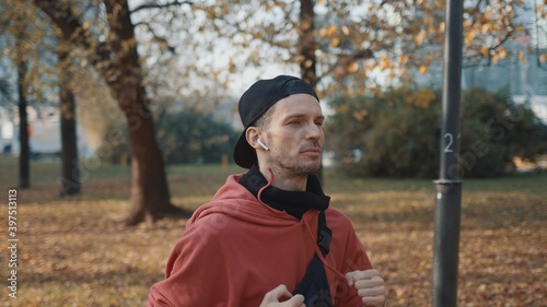 Man running in the park, young man in sport clothes and cap listening to music running through the park in the autumn day, middle shot, jogging concept © diproduction