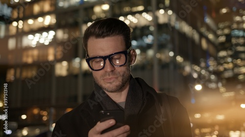 Man in eyeglasses with phone in hand on background of building with lights on. Gimbal night shot of caucasian man texting in phone near business building